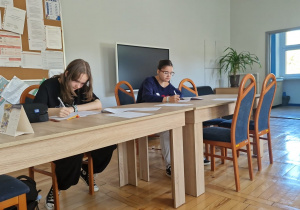 Dwie uczennice klasy ósmej siedzą przy długim, drewnianym stole w szkolnej sali. Pochylone nad kartkami papieru, samodzielnie rozwiązują zadania matematyczne. Na stole leżą arkusze z zadaniami i przybory do pisania.