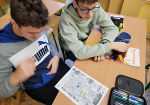 Troje uczniów siedzi przy szkolnej ławce i uczestniczy w matematycznej grze planszowej. Na stole leży papierowa plansza z polami zawierającymi zadania matematyczne oraz kolorowymi pionkami. Jeden z uczniów przesuwa rękę nad planszą, a pozostali obserwują przebieg gry.