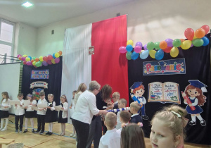 Sala gimnastyczna, uczniowie klasy pierwszej ustawieni w szeregu - pani dyrektor wielkim piórem pasuje uczniów, uczniowie otrzymują plakietki z logo SP40 oraz dyplomy